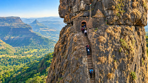 A dramatic mountain landscape with a tall rocky cliff, stone steps, and a small stone entrance where several hikers are walking. The valley below is covered in green vegetation and distant blue mountains are visible under a clear sky.