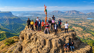 A group of hikers and climbers pose on a rocky mountain peak with a tall flag, overlooking a vast green valley and distant hills under a blue sky.