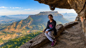 A woman sits on a rocky cliffside path overlooking a vast mountain landscape under a blue sky, framed by a natural rock overhang.