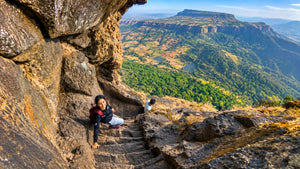 A scenic landscape photograph of a deep mountain valley with steep cliffs and green hills under a blue sky, with two people sitting on stone steps carved into a large rock formation in the foreground.